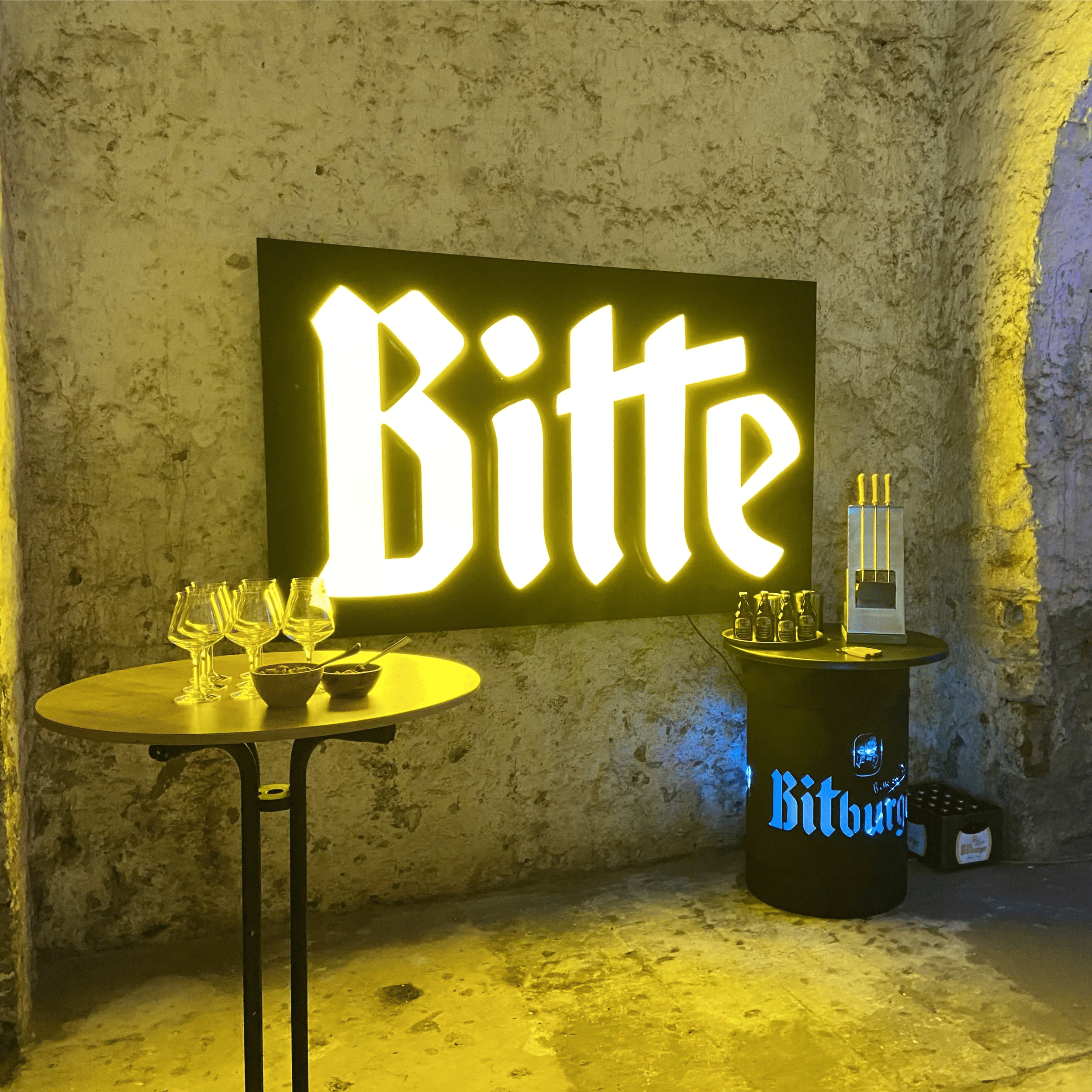 In einem Gewölbekeller mit warmen gelblichen Licht steht ein Stehtisch mit leeren Gläsern. rechts steht eine Bitburger Tonne mit Bierflaschen. An der Wand hängt ein großes beleuchtetes Werbeschild mit der Aufschrift "Bitte" im Bitburger Stil.