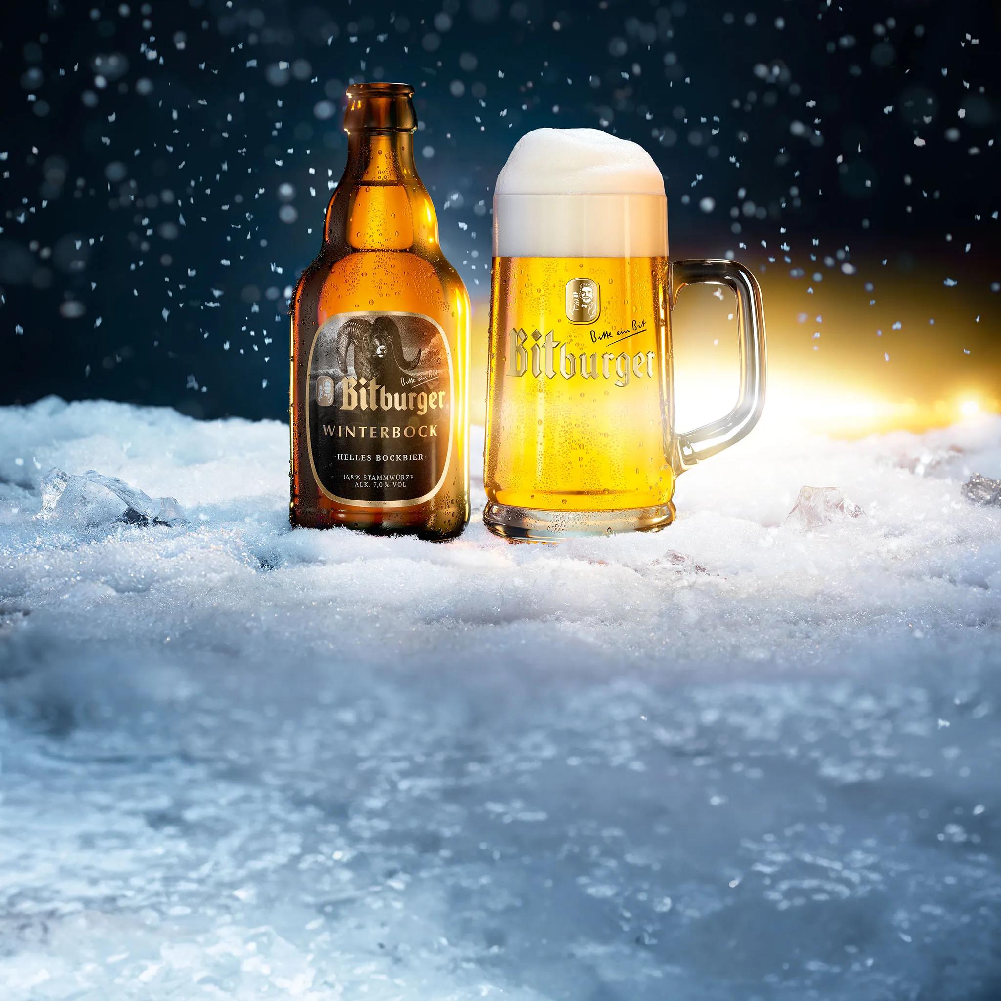 Eine Flasche Winterbock und ein Bierkrug stehen auf vereistem Schnee. Im Hintergrund ein dunkler Himmel und es schneit. Im Horizont ist ein gelber Lichtschein zu erkennen.